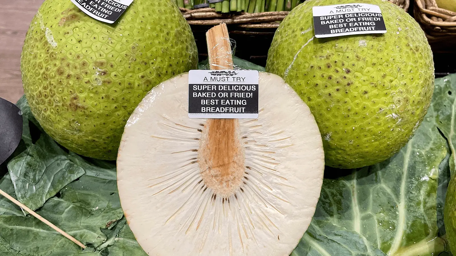 Close-up of two whole breadfruits with one sliced in half, displaying the white, textured interior. Labels read, "A Must Try! Super Delicious Baked or Fried! Best Eating Breadfruit." Green leafy vegetables surround the fruits, showcasing foods that define Hawaiian culture.