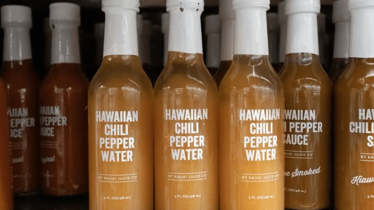 A shelf stocked with multiple bottles of Hawaiian Chili Pepper Water from Kauai Juice Co. Each bottle has a white cap and a label that reads "Hawaiian Chili Pepper Water" along with the brand's name and other details. This light amber condiment is one of the foods that define Hawaiian culture.