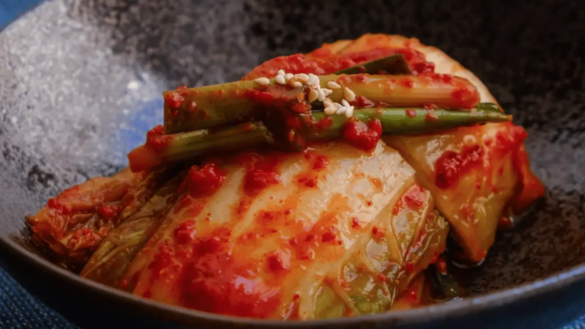 A close-up of a serving of kimchi in a dark bowl. The kimchi, one of the beloved condiments in many cultures, is garnished with sesame seeds and features a vibrant red chili paste coating, along with green onion stalks on top. The texture and layers of the cabbage are clearly visible.