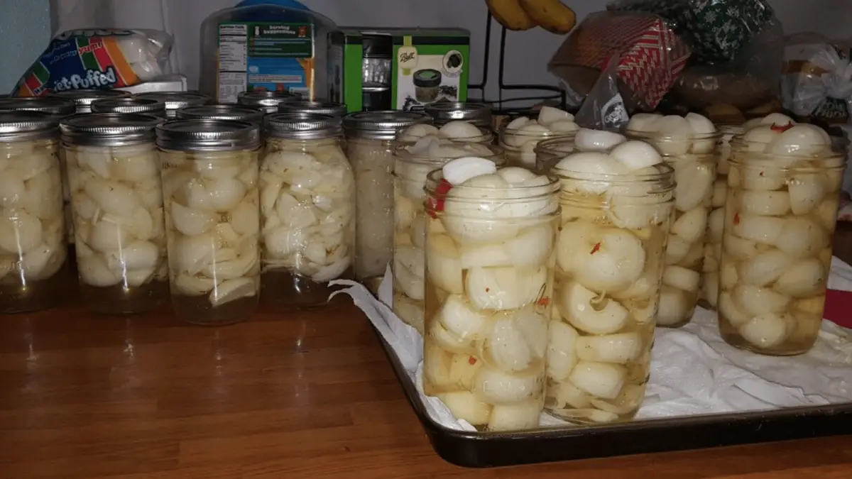 A collection of sealed jars filled with pickled onions on a wooden surface. Some jars are arranged on a tray lined with a cloth, while others are placed directly on the surface. Various kitchen items and food packages, including condiments, are visible in the background.