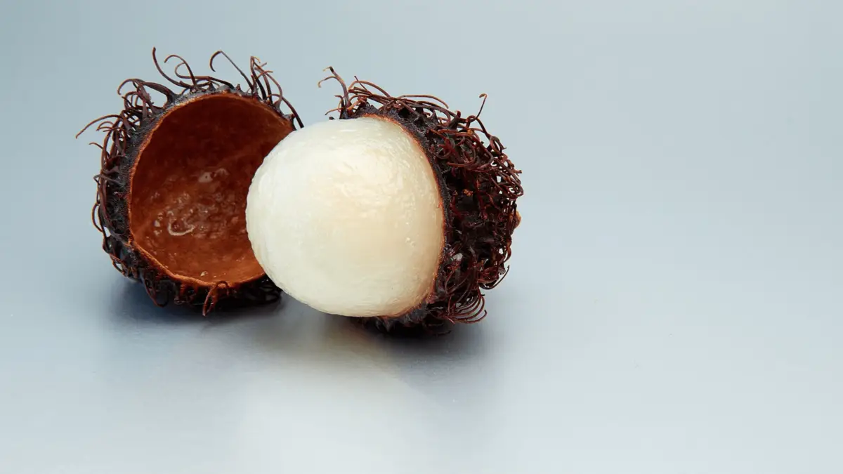 A rambutan with its hairy red-brown shell split open, showing the white, translucent flesh inside. The exotic fruit, a staple among foods that define Hawaiian culture, is set against a light gray background.