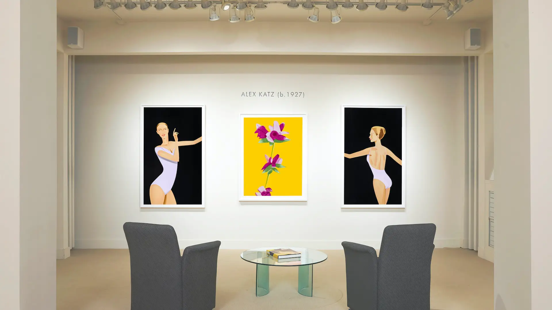 In one of the art galleries San Francisco boasts, a gallery room displays three artworks by Alex Katz: two portraits of women in white leotards on the left and right, and a vibrant, floral painting in the center. Two gray armchairs and a glass-top table with books are arranged in the foreground.