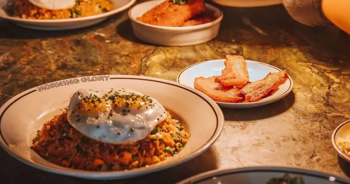 A plate of fried rice topped with two sunny-side-up eggs and garnished with chives is on the table. Beside it, there's a small plate of crispy bacon slices. The table has a rustic appearance, and there are other dishes partially visible in the background.