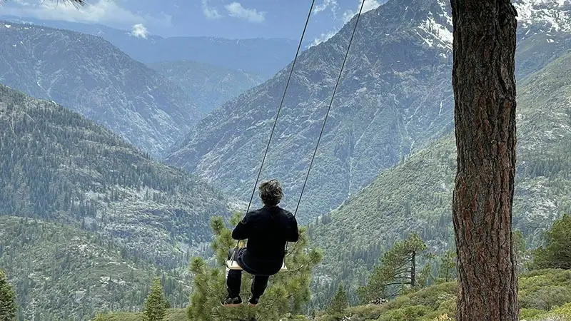 A person is sitting on a swing attached to a tall tree, overlooking the picturesque Tahoe mountain landscape. The mountains are covered in greenery with some patches of snow, and the sky is partly cloudy. The scene evokes a sense of peace and natural beauty.