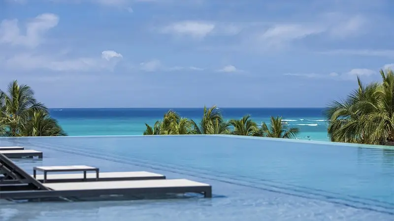 A serene infinity pool with lounge chairs on the left overlooks a turquoise sea. Palm trees line the horizon under a partly cloudy sky, creating a tranquil and picturesque tropical beach scene often found at the best hotels on Oahu.