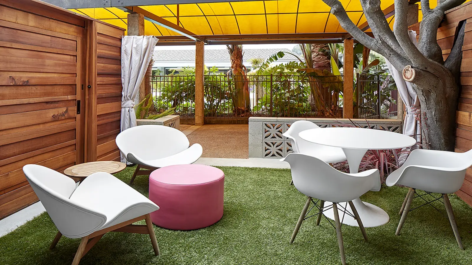 A cozy outdoor patio on the Monterey Peninsula features modern white chairs, a round white table, and a circular pink ottoman on green artificial grass. The area is shaded by a yellow awning and bordered by a wooden fence and lush tropical plants. A tree grows through the wooden deck—an ideal spot for those seeking where to stay.