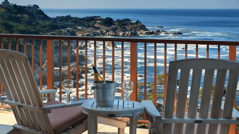 Two wooden chairs face each other on a balcony of one of the most romantic hotels in the Bay Area, overlooking a scenic rocky coastline and the ocean. Between the chairs, there's a small table with a chilled bottle of wine in an ice bucket and two empty glasses. The sky is clear and sunny.