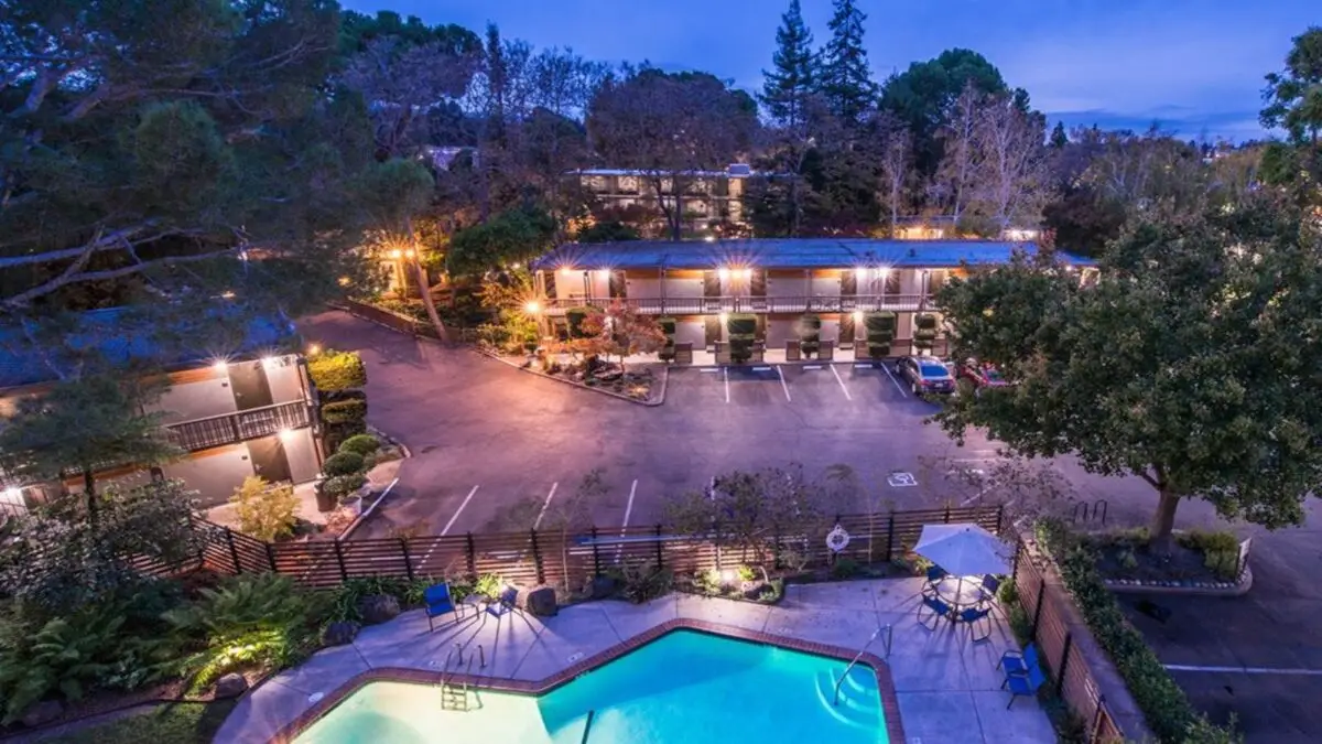 A twilight view of the quiet Creekside Inn reveals a softly lit exterior. The scene includes a glowing swimming pool in the foreground, surrounded by lounge chairs and a table with an umbrella. Trees and bushes border the well-maintained property, with cars parked in front of the rooms.