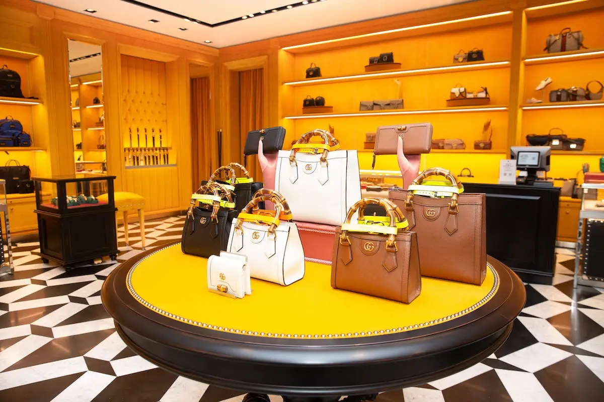 A display of luxury handbags in various colors, including black, white, brown, and two-tone designs, graces a round table with a yellow surface at DFS's high-end boutique. The background features shelves filled with more bags and accessories against a vibrant yellow backdrop.