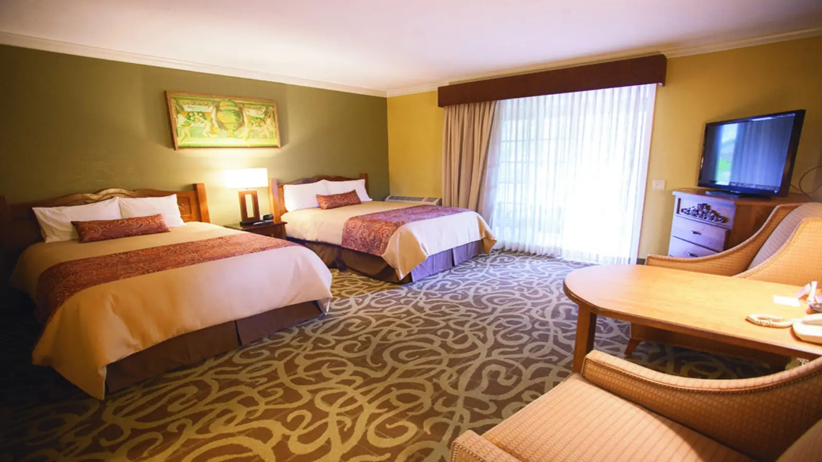 A well-lit hotel room at one of the best value hotels in Napa Valley features two double beds with decorative pillows and blankets. The room includes a wooden table with two cushioned chairs, a large window with floor-to-ceiling curtains, a flat-screen TV on a dresser, and a patterned carpet. A painting hangs on the wall.