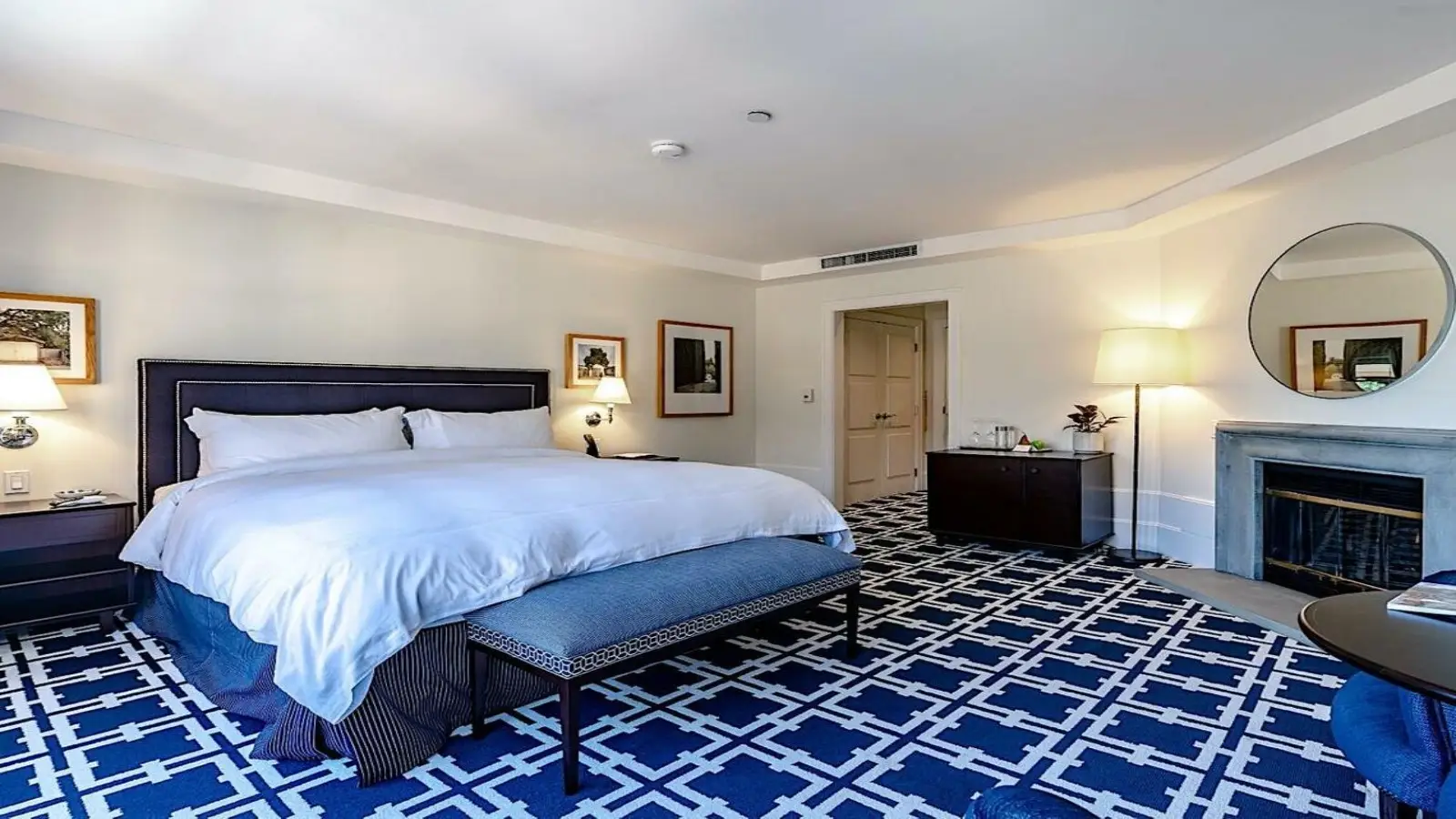 A spacious hotel room at El Prado Hotel features a king-sized bed with white linens, a patterned blue carpet, a fireplace, a large round mirror above a wooden dresser, two wall art pieces, a cozy reading nook with a lamp, and a doorway leading to an adjacent area.