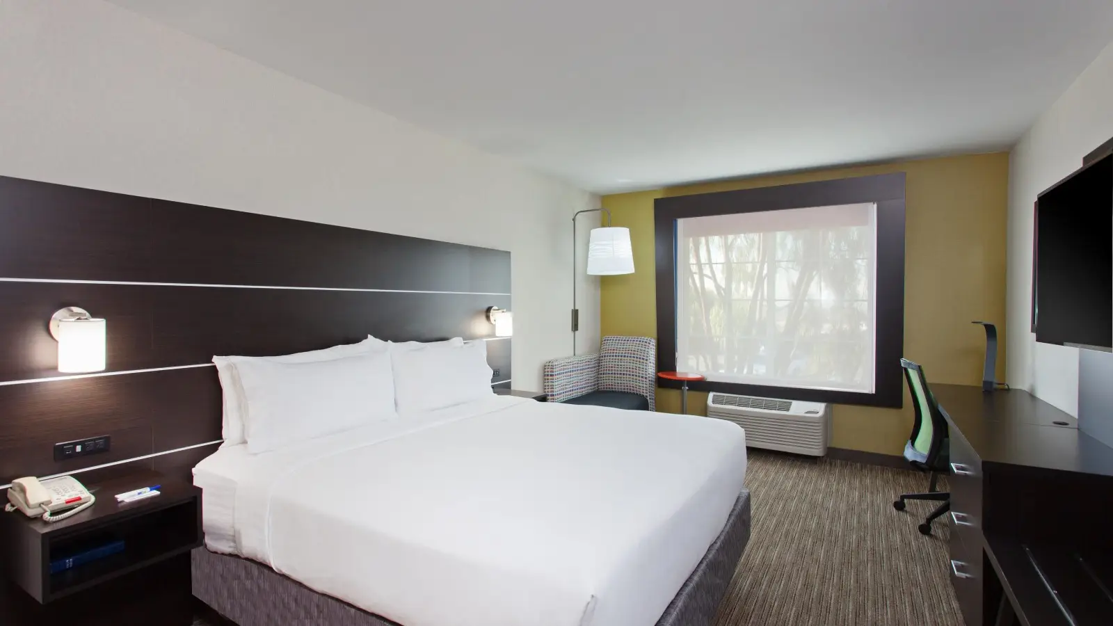 A modern Holiday Inn Express Oakland Airport hotel room features a king-sized bed with white linens, a dark headboard with wall-mounted lights, a large window with a pull-down shade, a cushioned chair, a desk with a rolling chair, and a flatscreen TV mounted on the wall.
