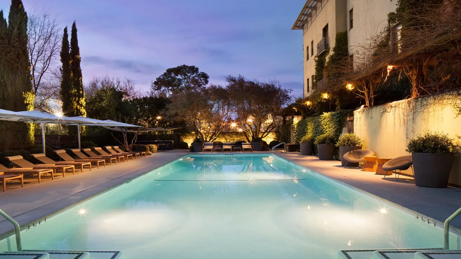 A serene outdoor pool scene at dusk with lounge chairs lining both sides, large potted plants, and umbrellas for shade on the left. Trees and string lights add ambiance. A modern building in the background enhances the elegant atmosphere—an ideal spot when considering where to stay in Sonoma.