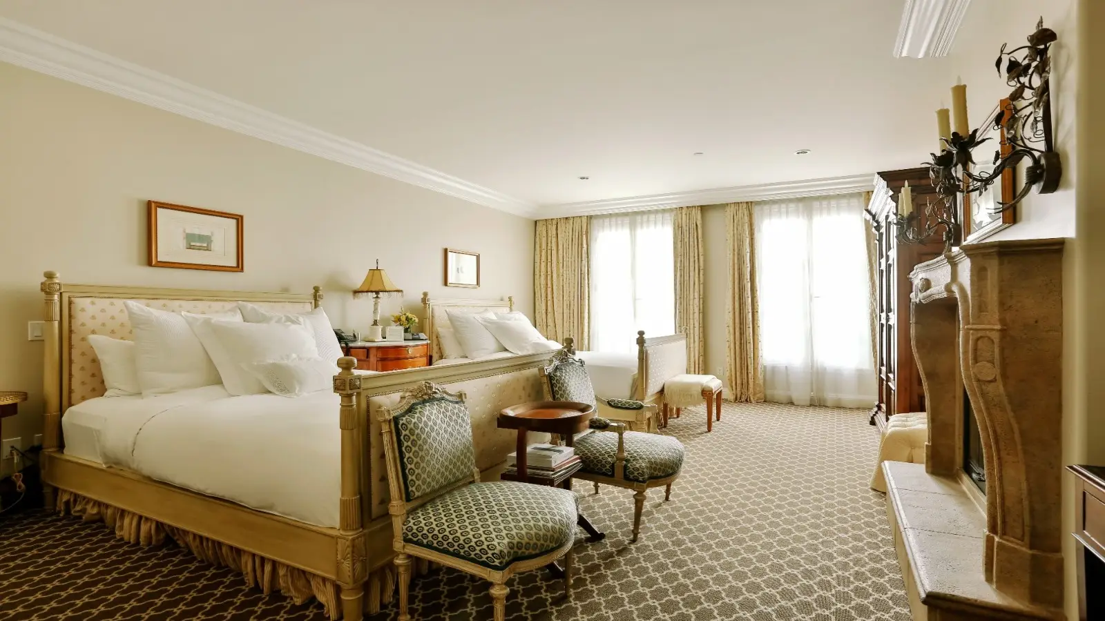 A well-lit hotel room where to stay in Sonoma, featuring a large bed with white linens, cushioned seating at the foot, and a patterned carpet. There's a fireplace on the right, chairs and a table in the center, and tall windows with light curtains in the background.