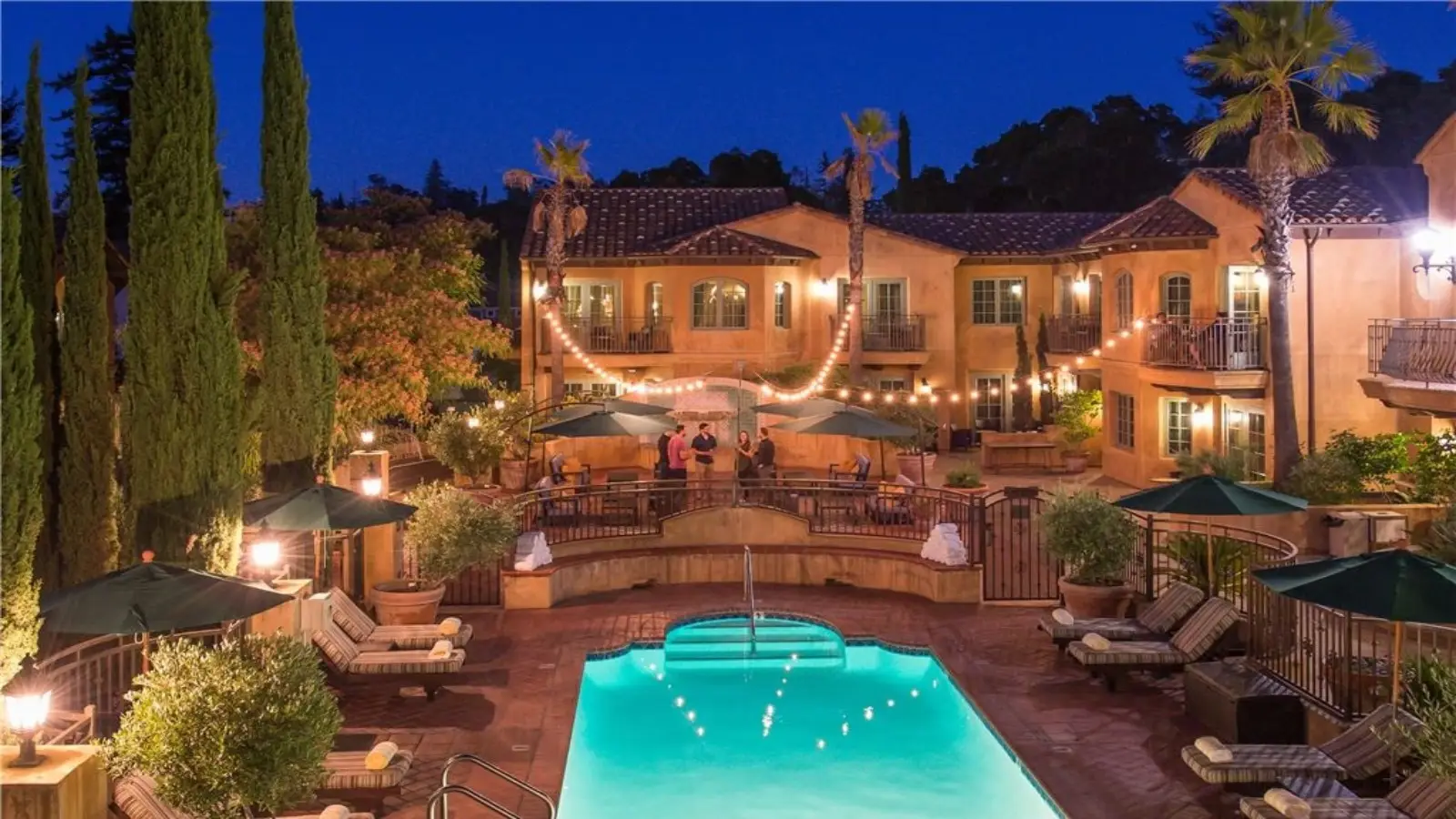 A cozy, well-lit courtyard at Hotel Los Gatos features a glistening swimming pool surrounded by lounge chairs and umbrellas. String lights hang above the scene, illuminating a two-story building with balconies set against a twilight sky. Palm trees and lush greenery enhance the serene ambiance.