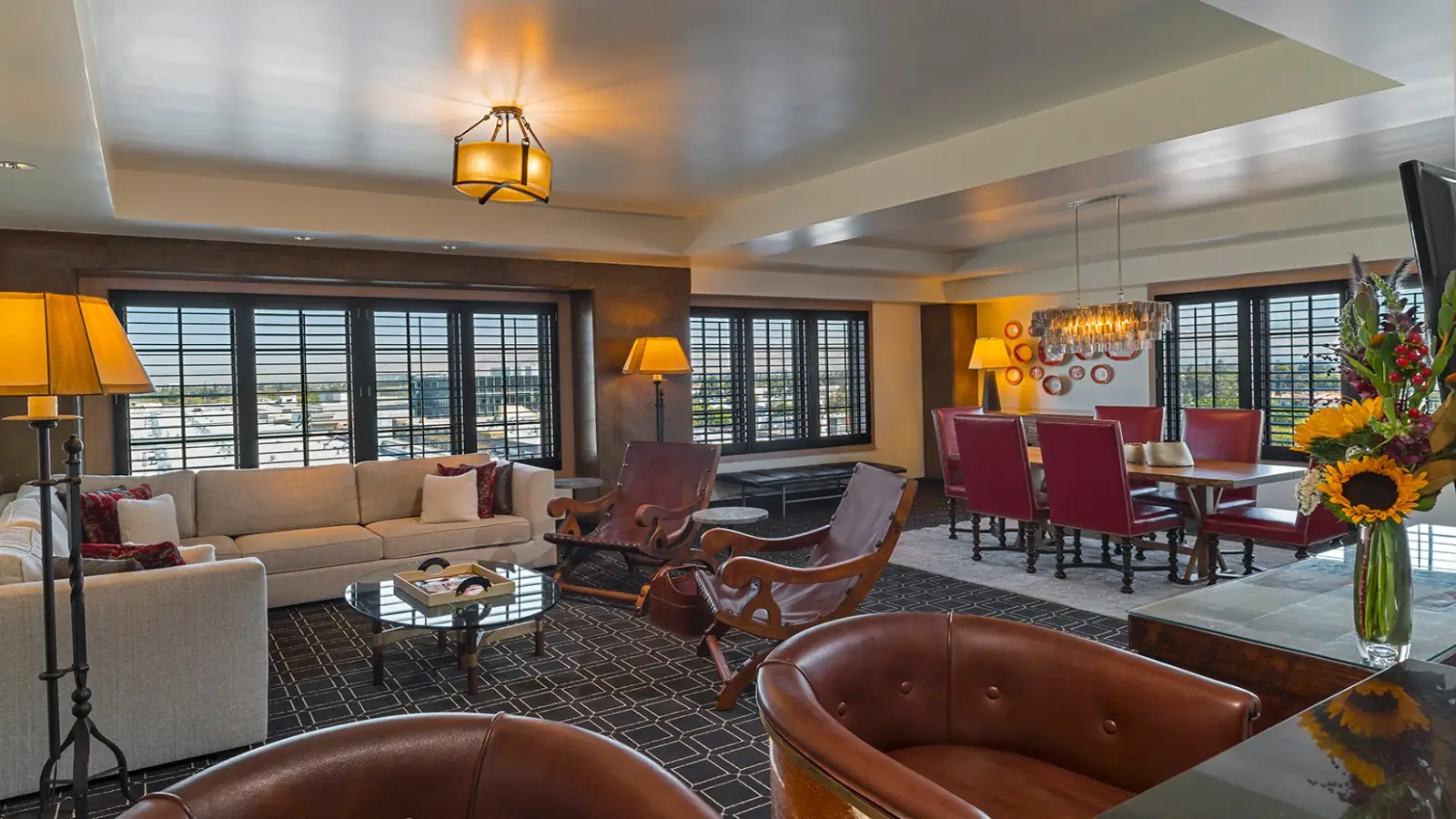A luxurious living and dining area with contemporary decor reminiscent of Hotel Valencia Santana Row. It features a beige sectional sofa, wooden chairs, a glass coffee table, a dining table with red leather chairs, and large windows with black frames. There are modern light fixtures, lamps, and floral arrangements.