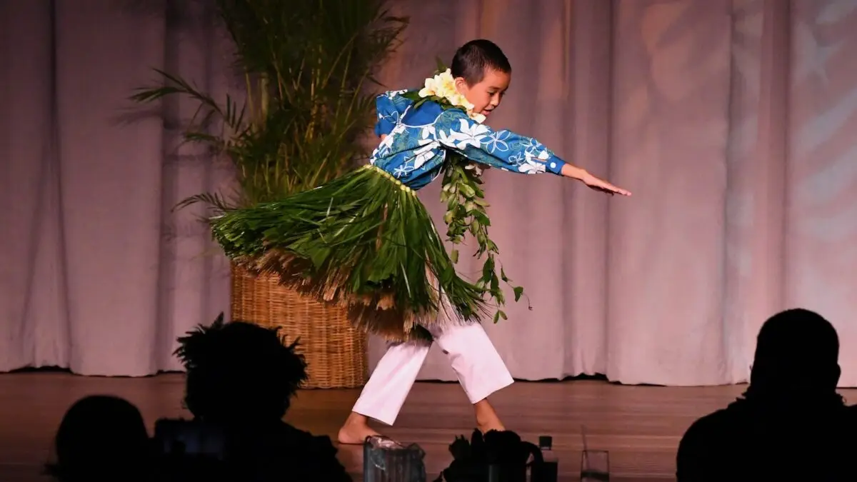 A young boy in traditional Hawaiian attire, including a grass skirt and floral leis, performs a hula dance on stage at the Kā‘anapali Beach Hotel. He extends one arm forward while bending his knees. Potted greenery and a curtain backdrop are visible, and the silhouettes of the audience can be seen.