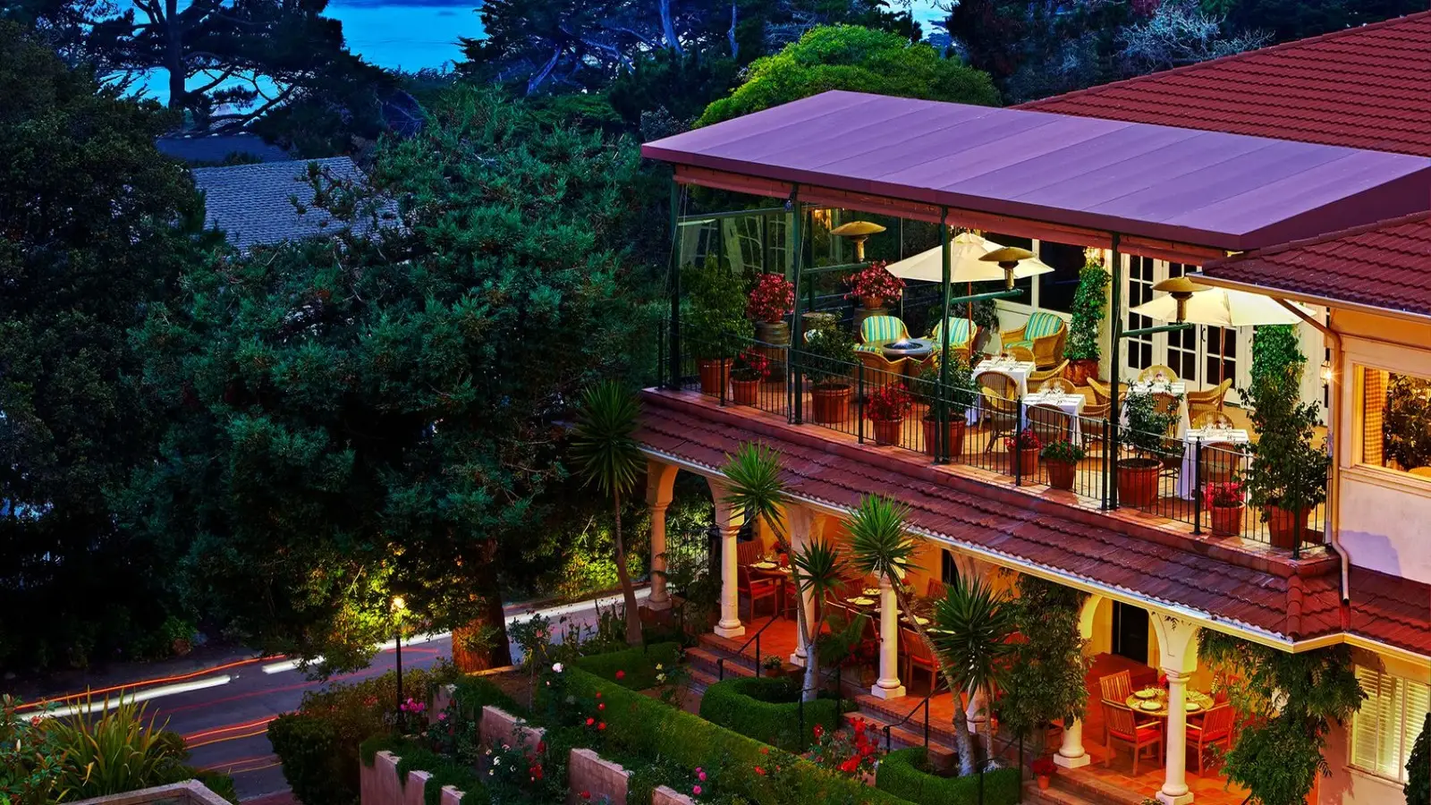 A two-story building with a glass-enclosed dining area on the upper floor, featuring tables and chairs under hanging umbrellas offers a perfect getaway where to stay on the Monterey Peninsula. The surrounding area is lush with greenery, palm trees, and bright flowers, while the background reveals a view of the ocean at dusk.