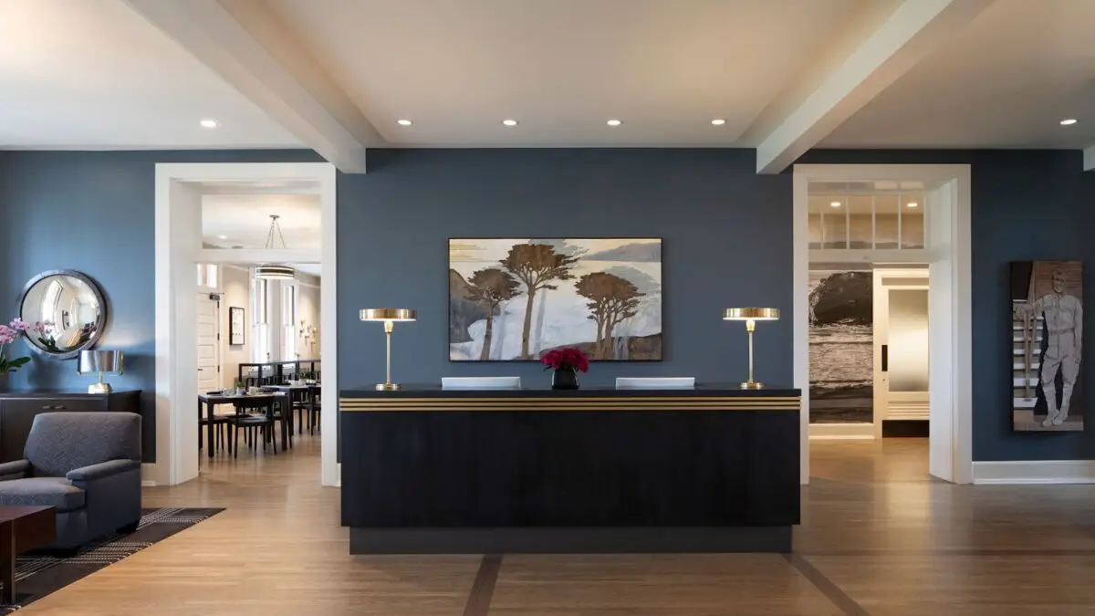 A modern reception area at the Lodge at the Presidio features a dark wooden counter topped with two sleek desk lamps and flower arrangements. Behind the counter hangs a landscape painting. The walls are painted a deep blue, with adjacent rooms visible in the background.