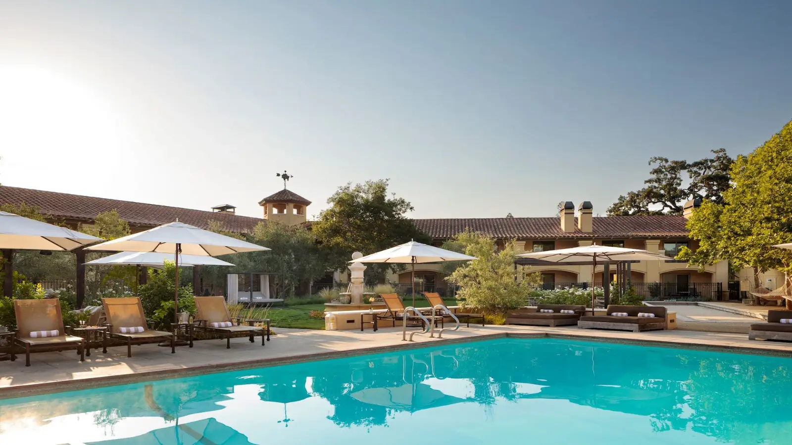 Outdoor pool area at one of the best value hotels in Napa Valley, complete with lounge chairs and umbrellas arranged around the pool. The resort building features a tile roof and arched windows, while lush trees and landscaping provide an inviting atmosphere. The sun shines brightly, creating a serene ambiance.