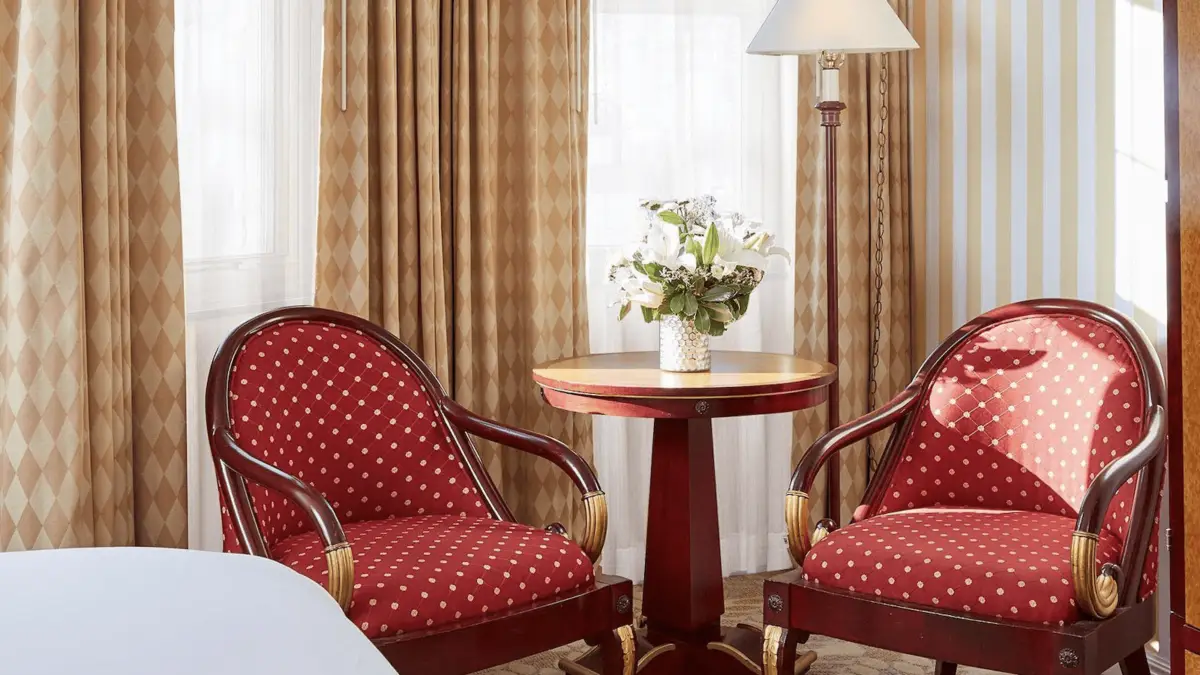 A cozy sitting area at the Orchard Hotel features two red armchairs with white polka dots, placed around a round wooden table with a floral arrangement in a vase. Beige curtains with a diamond pattern and a floor lamp with a white shade complete the serene and inviting atmosphere.