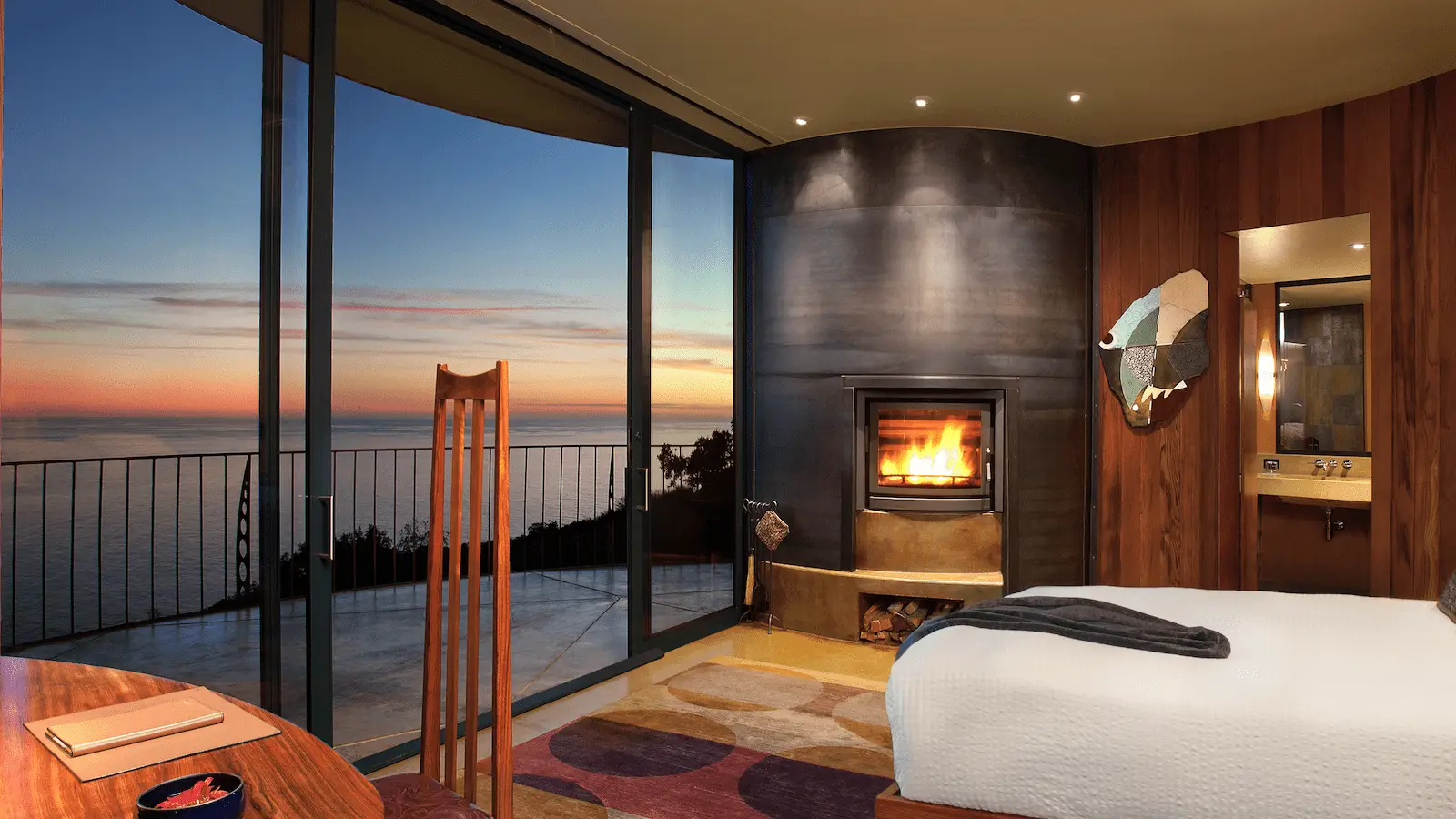 A cozy bedroom with a modern design featuring a fireplace and a large curved floor-to-ceiling window offering an ocean view. The room includes a bed, a wooden chair, and a desk, with a colorful rug on the floor. If you're wondering where to stay in Monterey Peninsula, this room at sunset is perfect.