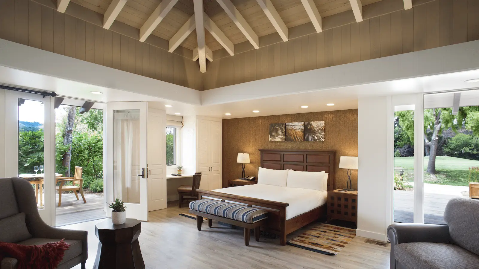 A spacious, cozy hotel room with wooden ceilings and a large, comfortable bed. The room features modern furniture, a desk, an armchair, and large windows opening to a lush garden at the renowned Quail Lodge Golf Club. A small seating area is visible outside on a patio.