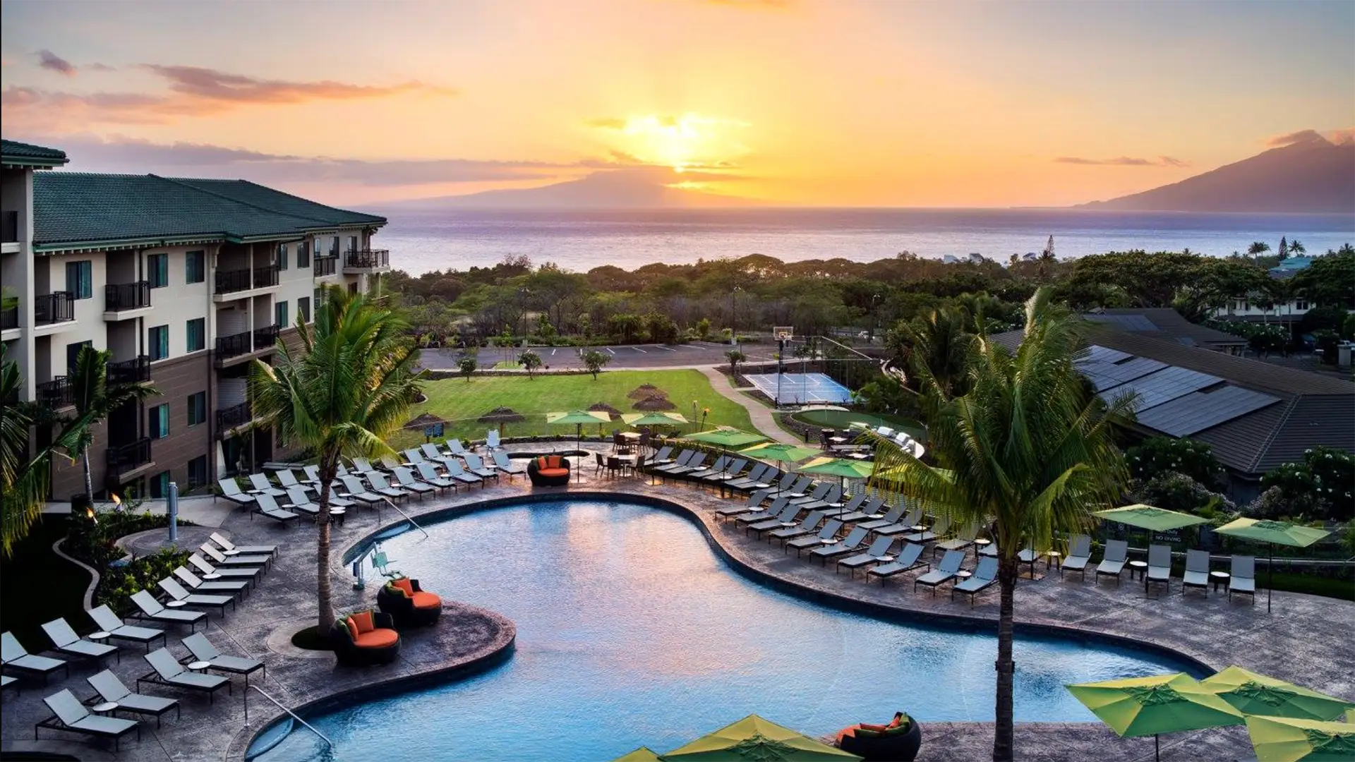 Sunset view of a luxurious resort featuring a large, curvy swimming pool surrounded by lounge chairs and palm trees. The resort overlooks a scenic landscape with lush greenery and the ocean in the distance, with a colorful sky at dusk adding to the tranquil atmosphere—truly one of the best places for those wondering where to stay in Wailes.