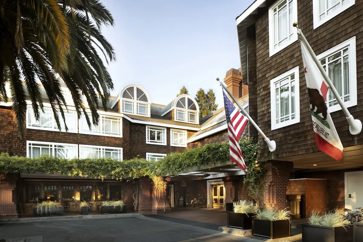 The image shows the exterior of Stanford Park Hotel with brown brick walls covered in greenery. The building features large windows and arched details on the roof. Two flags, one American and one Californian, stand at the entrance, while palm trees and additional greenery enhance the surroundings.