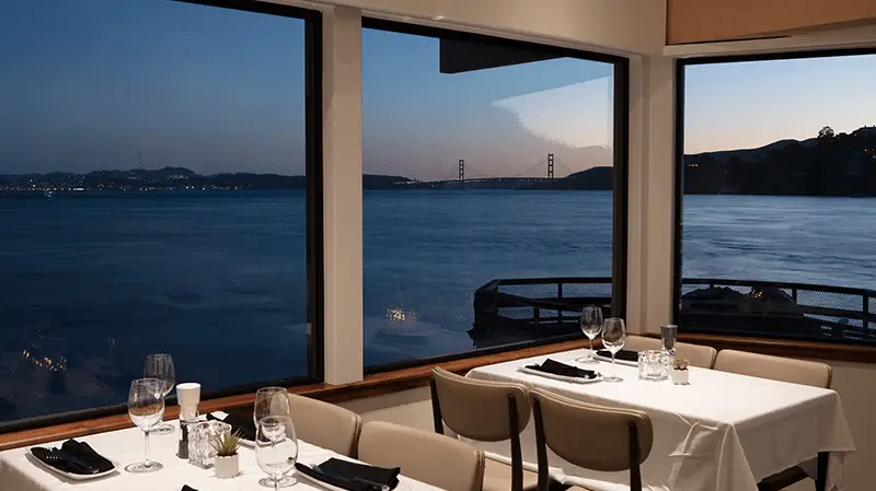 A cozy restaurant with large windows overlooking a serene body of water and a bridge in the distance during sunset. Tables are set with wine glasses and napkins, ready for diners to enjoy the picturesque view—a hidden gem when deciding where to eat in the Bay Area.