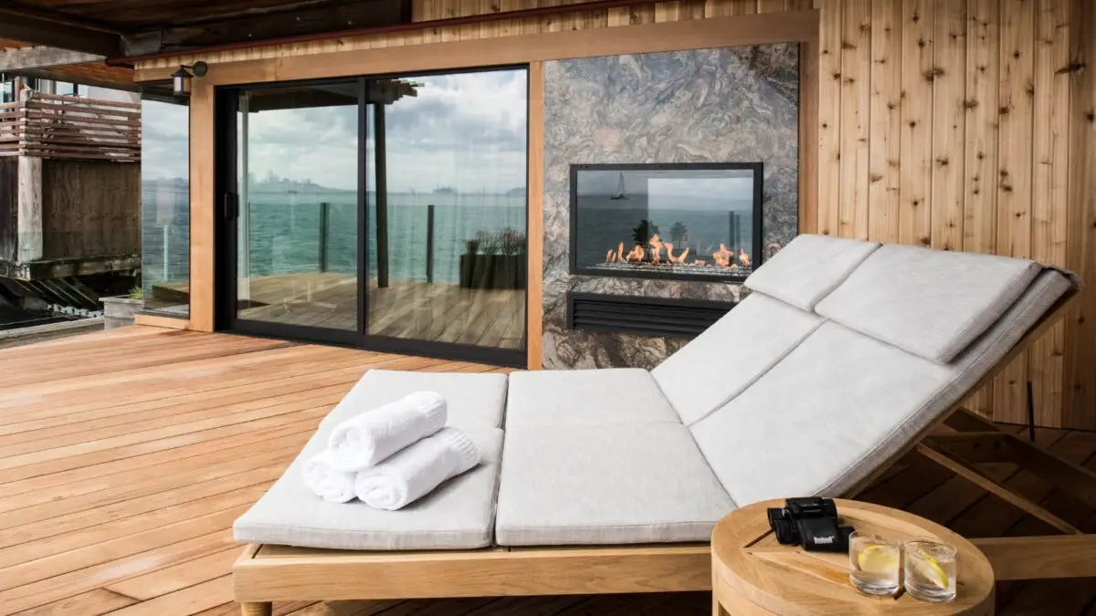 A luxurious outdoor deck at the Inn Above Tide features a cushioned double chaise lounge with white towels on it, situated beside a small wooden round table holding two drinks. The deck overlooks the ocean and includes a sliding glass door and an outdoor fireplace.