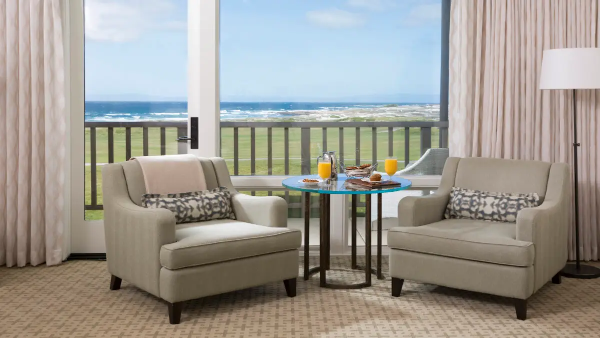 Two beige armchairs with patterned cushions face a small round table topped with breakfast items, including orange juice and pastries. The room at the Inn at Spanish Bay features a large window with a view of a green lawn and the ocean in the distance. Light curtains frame the window.