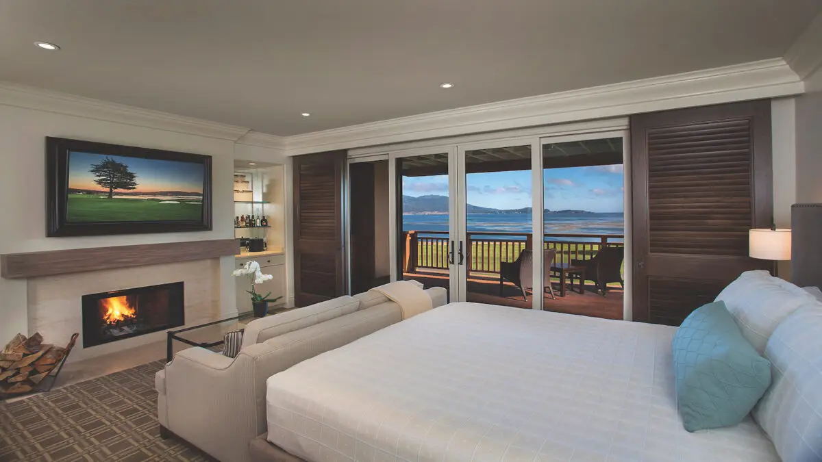 A cozy bedroom at the Lodge at Pebble Beach features a large bed with white linens, a fireplace with a fire burning, and a wall-mounted TV. Sliding glass doors open to a balcony that offers a scenic view of the lake or ocean and distant mountains. A small seating area and bar are present.