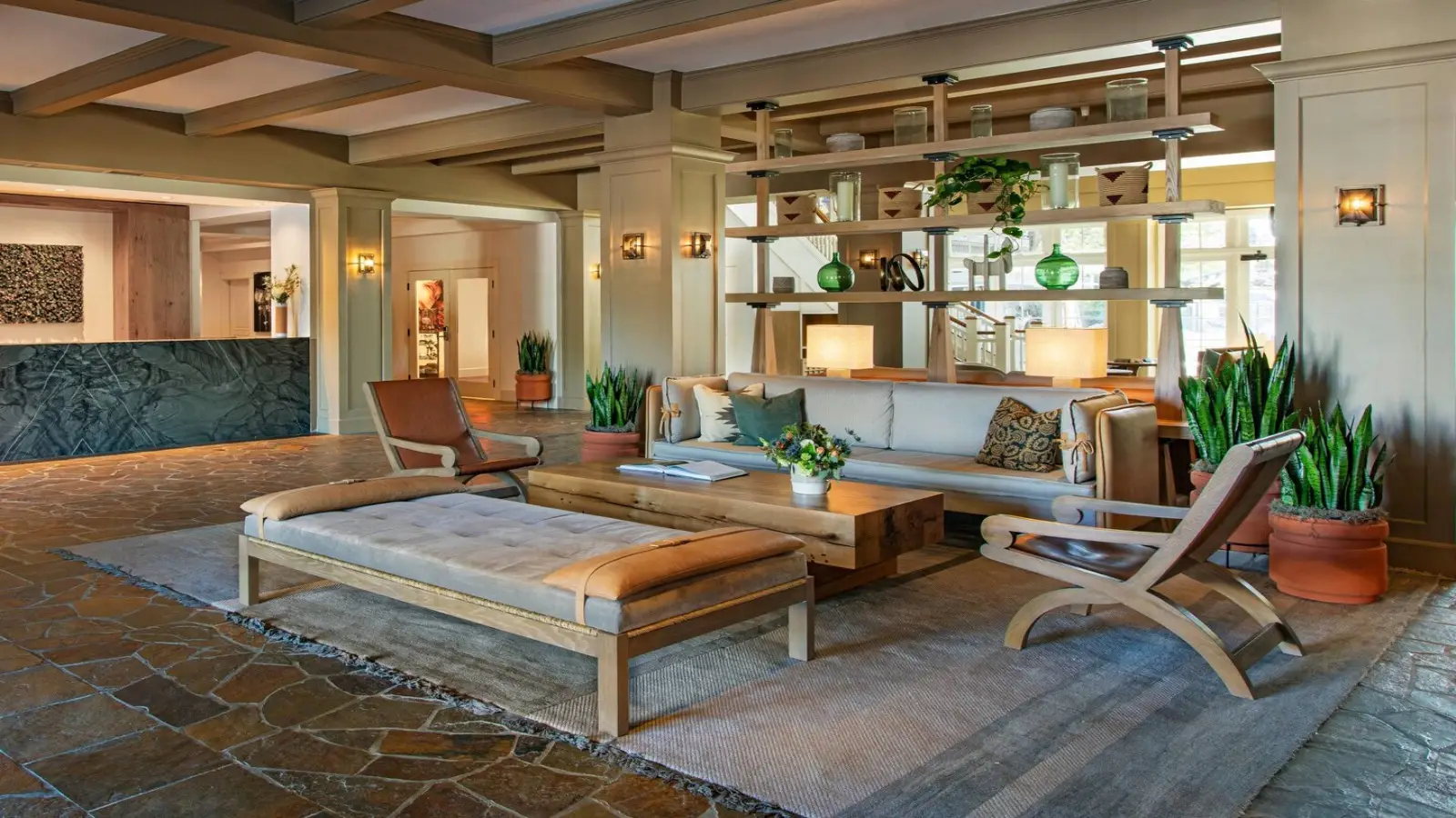 A cozy hotel lobby, perfect for those deciding where to stay in Sonoma, features a seating area with a wooden coffee table, beige cushioned chairs, a beige daybed, and potted plants. Behind the seating is a decorative shelving unit with vases and lamps. The reception desk with a marble front is seen in the background.