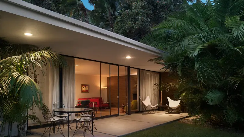 A modern, single-story house in Palm Springs with large glass sliding doors opening to a patio. The patio features a round table with wire chairs and two white lounge chairs. The house is surrounded by lush greenery, illuminated by soft outdoor lighting, making it an ideal retreat for those seeking where to stay in Palm Springs.