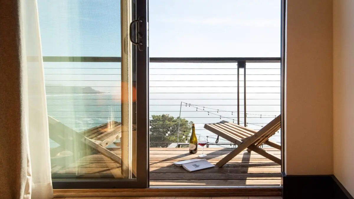 Discover a cozy balcony at your next where-to-stay North Bay spot, offering a panoramic sea view. It features two wooden lounge chairs, a wine bottle, a glass of red wine, and a book on a small table. Soft sunlight filters through the sliding glass door adorned with light curtains, and string lights hang along the railing.