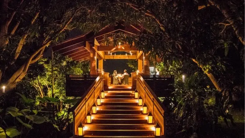 A warmly lit wooden staircase leads to a cozy, illuminated dining area surrounded by lush greenery and trees. Two people are seated at a table under a wooden canopy, creating a romantic, serene atmosphere in the night. Lanterns line the stairs, adding to the ambiance of this perfect romantic getaway in Hawaii.