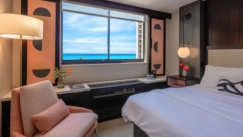 A stylish hotel room with a window view of the ocean. The room features a bed with white linens, a pink chair with an orange pillow, and a bedside table with a small plant and phone. A large floor lamp is next to the chair, and the walls have decorative accents, making it one of the best hotels on Oahu.