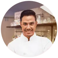 A smiling person in a white chef's coat stands in a kitchen at Ghirardelli Square. The background includes shelves with glassware and various kitchen utensils. The image is circular in shape with a soft, blurred edge effect.