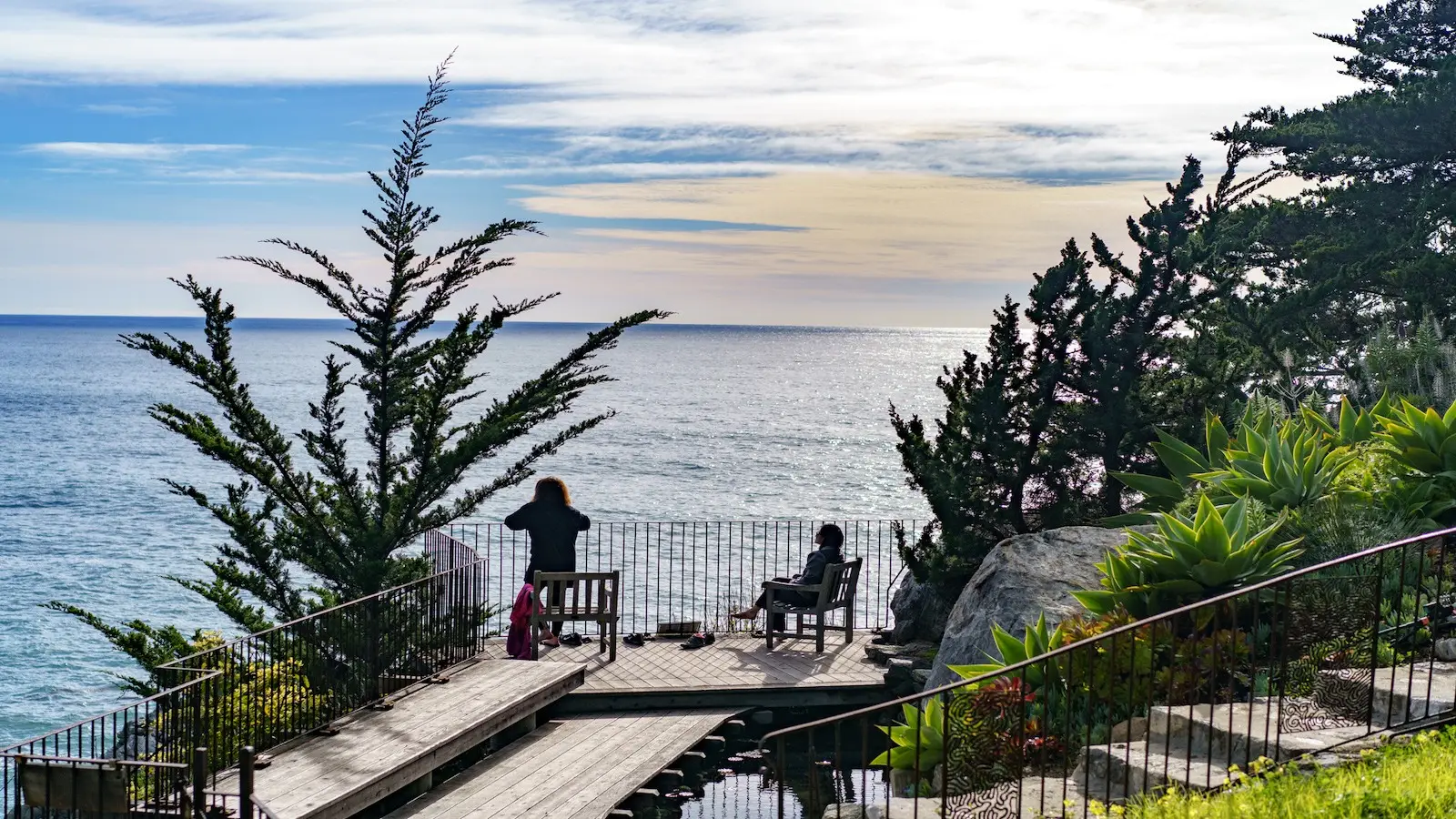 Two people enjoy a scenic view from a wooden deck overlooking a vast ocean, a perfect spot for local getaways. One stands, capturing the moment with their phone, while the other relaxes on a bench. The deck is surrounded by trees and green foliage, with a dramatic coastline and blue sky in the background.