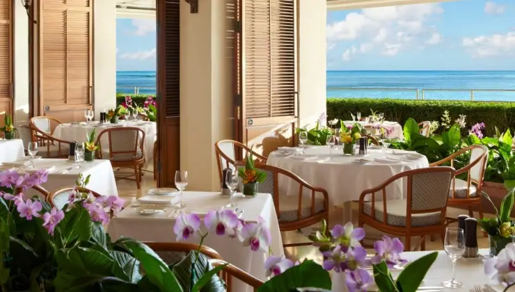 An elegant seaside restaurant with wooden chairs and white tablecloth-covered tables, adorned with floral centerpieces. The open wooden shutters offer a view of the blue ocean and sky. Lush green plants and blooming orchids add a tropical ambiance to the setting, rivaling the best hotels on Oahu.