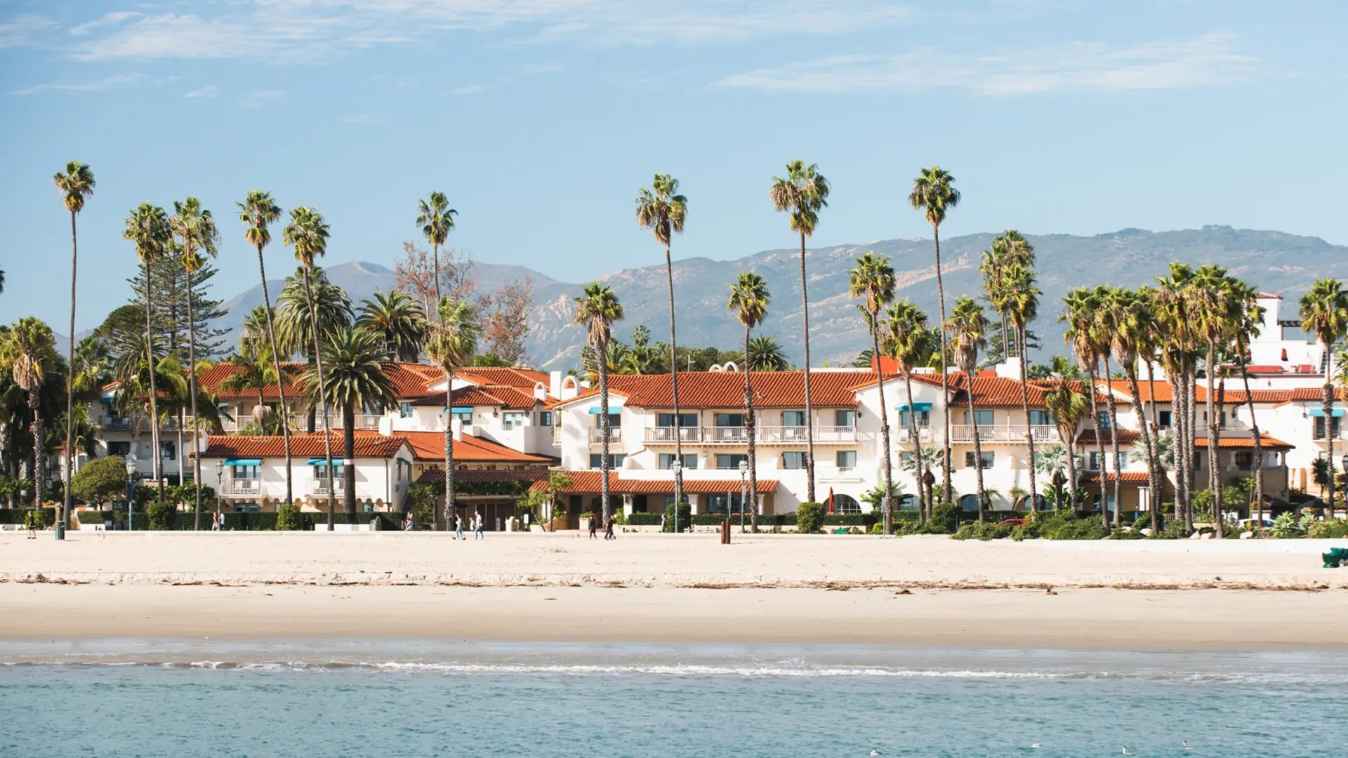 A scenic view of a beachfront resort with white buildings and red-tiled roofs, surrounded by tall palm trees. In the background, mountains are visible under a clear blue sky. The sandy beach and calm ocean water appear in the foreground, evoking the charm of Santa Barbara hotels.