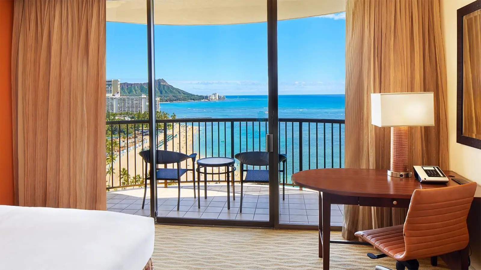 A hotel room at one of the best hotels on Oahu features a cozy bed in the foreground. A desk with a lamp and chair sits beside the sliding glass doors. Through the open doors, a balcony overlooks a scenic ocean view with a distant mountain under a clear blue sky. Two chairs and a table are on the balcony.