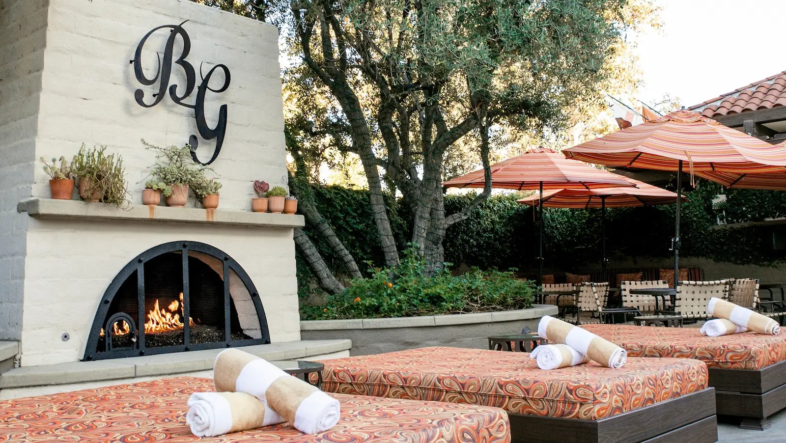 Outdoor lounge area with patterned lounge chairs, rolled towels, and large umbrellas. A fireplace with a "BG" monogram has potted plants arranged on the mantle. Trees and shaded seating are in the background, creating a cozy, inviting atmosphere—perfect for anyone wondering where to stay in Palm Springs.