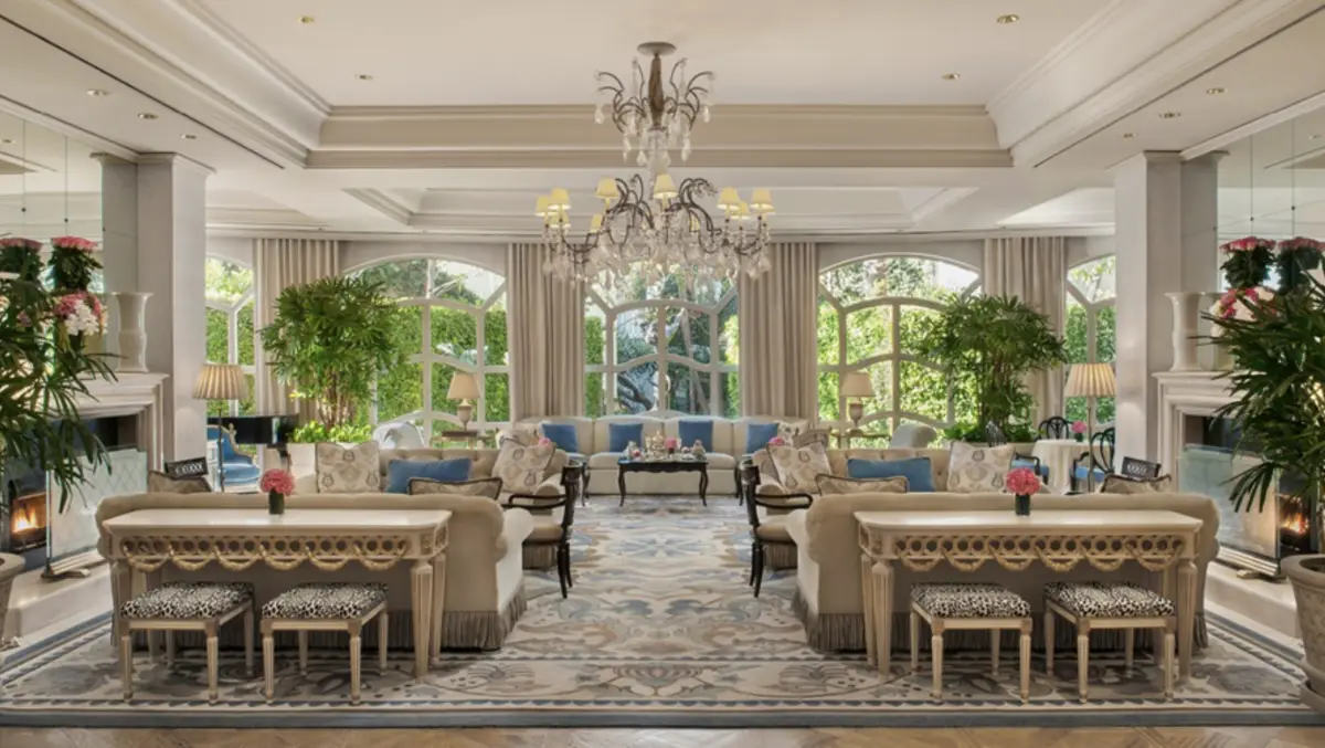 A spacious, luxurious living room at The Peninsula Beverly Hills boasts large arched windows allowing natural light to fill the space. It features elegant chandeliers, plush sofas, cushioned benches, and potted plants. The room is decorated with neutral tones and intricate patterned rugs.