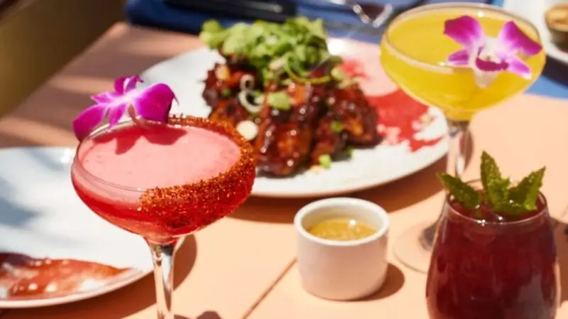 Close-up of a table with vibrant cocktails garnished with flowers and mint. There is a red drink in a glass with a chili rim, a yellow drink, and a deep red drink in front. In the background, where to stay Palm Springs recommendations are discussed over a plate of food garnished with greens.