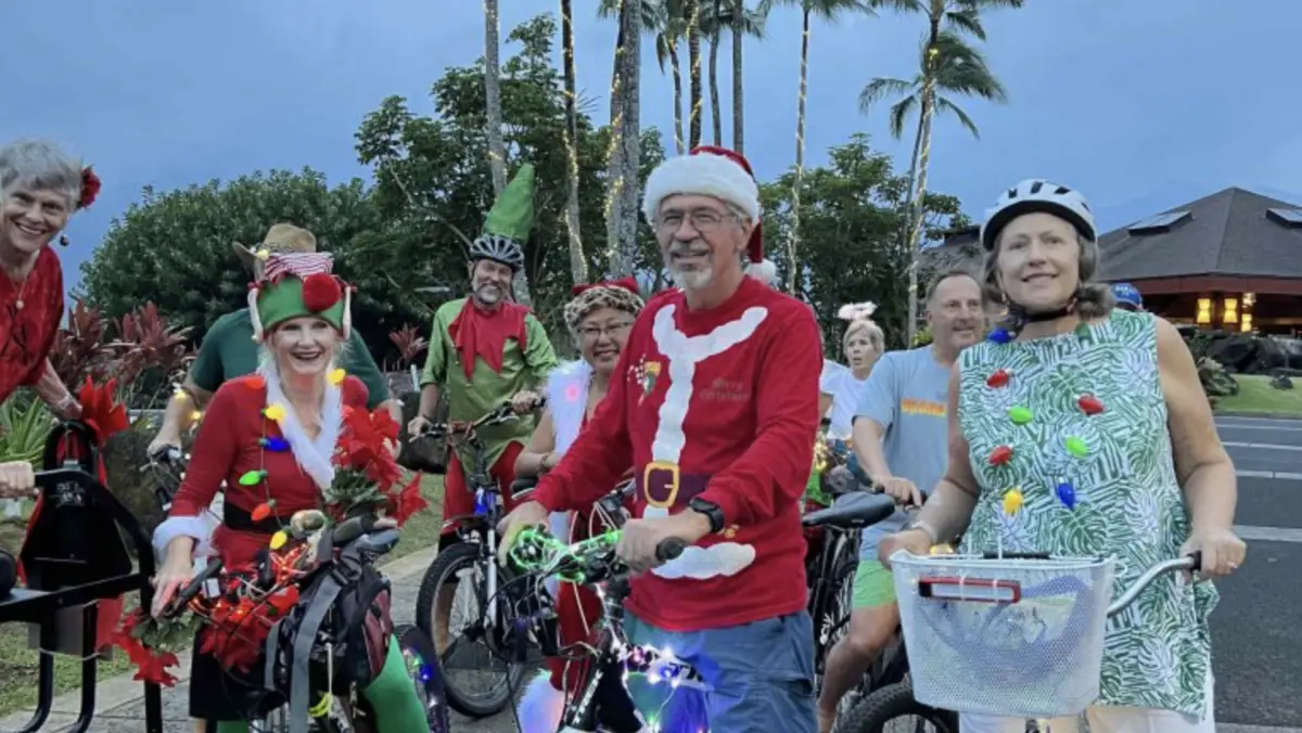 A group of people in festive holiday attire, including a Santa costume and elf outfits, are standing with bicycles. The bikes are adorned with lights and decorations. Palm trees and a small hut are in the background, capturing the essence of Princeville Kauai Annual Events in a tropical setting.