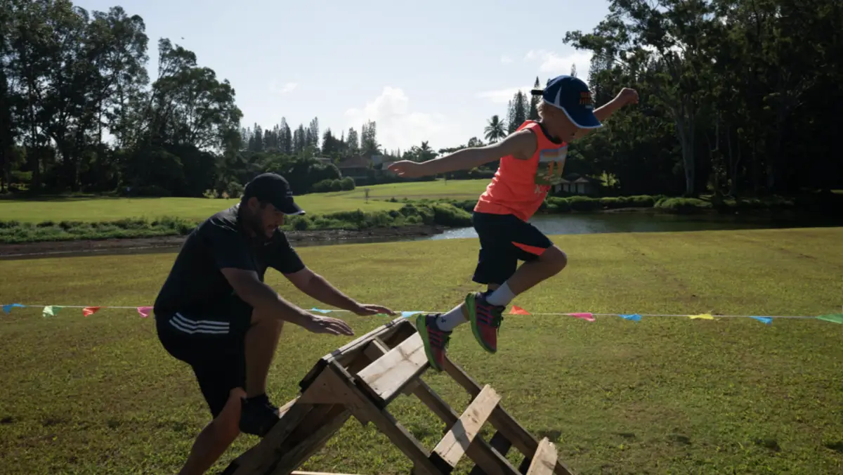 A man and a child participate in an outdoor obstacle course at one of the Princeville Kauai Annual Events. The child, wearing an orange shirt, blue cap, and sneakers, is jumping off a wooden structure while the man in black athletic wear climbs behind him. Green grass and trees are in the background.