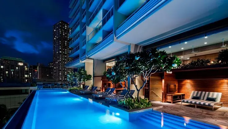 Night view of an illuminated rooftop infinity pool at one of the best hotels on Oahu. Tables and lounge chairs are arranged poolside under trees. Tall buildings are visible in the background, and the hotel interior can be seen through large glass windows.