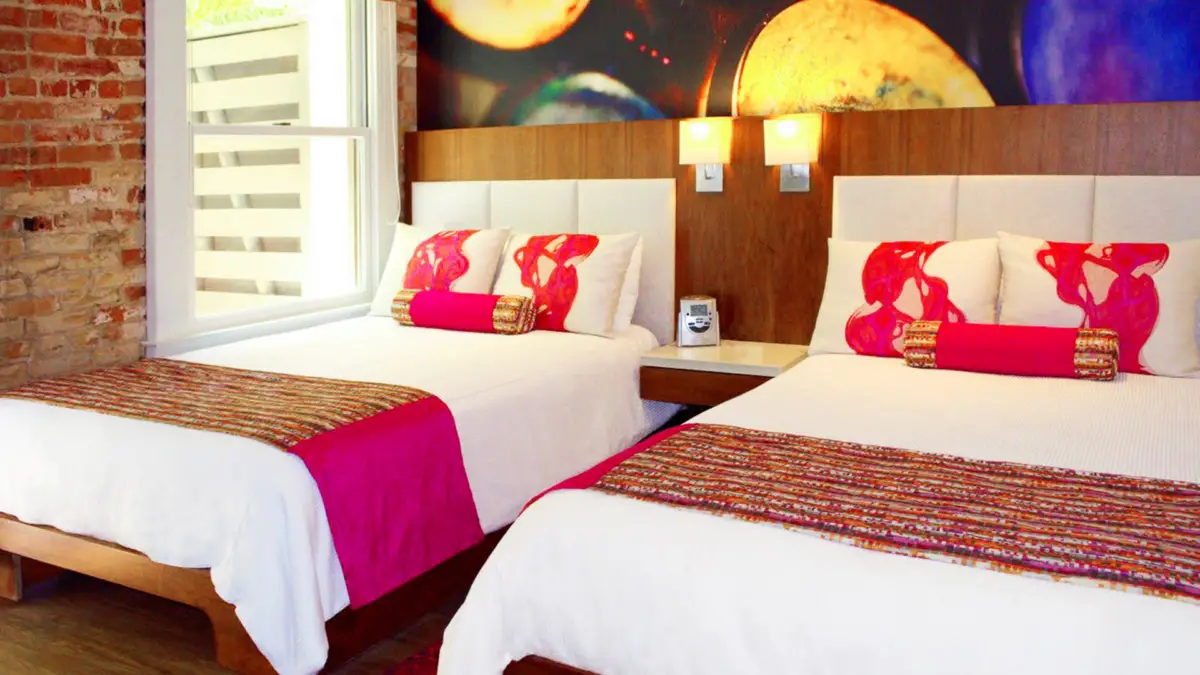 A bright and colorful hotel room, perfect for those considering where to stay in Santa Barbara, features two double beds adorned with white linens, vibrant pink pillows, and runners. A nightstand with a phone and lamp sits in between, while a space-themed mural with planets decorates the wall. A large window graces one side.